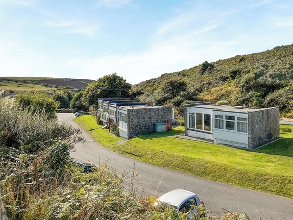 Une rangée de maisons mobiles modernes et compactes, dotées de façades en pierre et de grandes fenêtres, est située dans une vallée verdoyante et luxuriante, sous un ciel dégagé.