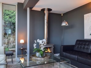 A modern living room with a glass coffee table, a black leather sofa, a fireplace, and a view of greenery outside.