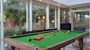 Una mesa de billar con bolas, una silla de madera y un televisor en una habitación con ventanas grandes.