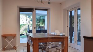 Un comedor con mesa de madera, sillas de plástico transparente, ventanas amplias y una araña.