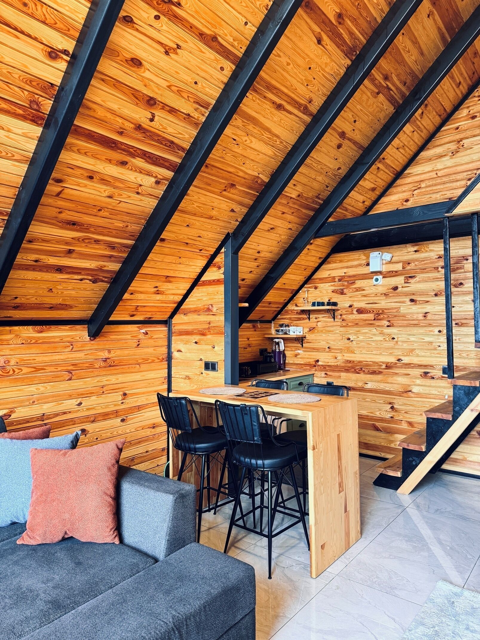 Quarto moderno no estilo loft, com vigas de madeira aparentes e teto inclinado. Há uma sala de jantar com mesa e cadeiras de madeira, um sofá com almofadas, e uma cozinha com eletrodomésticos.