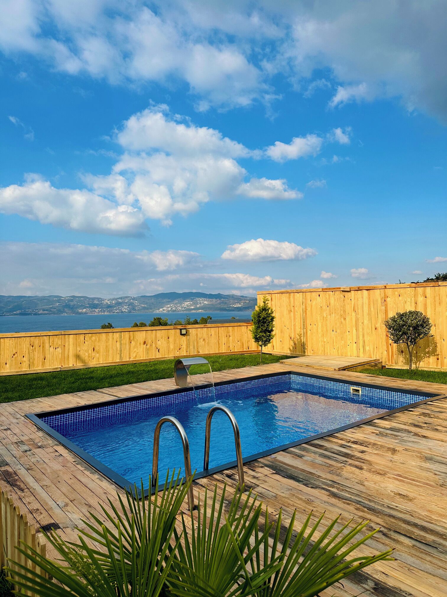 A rectangular swimming pool with a wooden deck, surrounded by a wooden fence, and a view of distant mountains and a body of water.