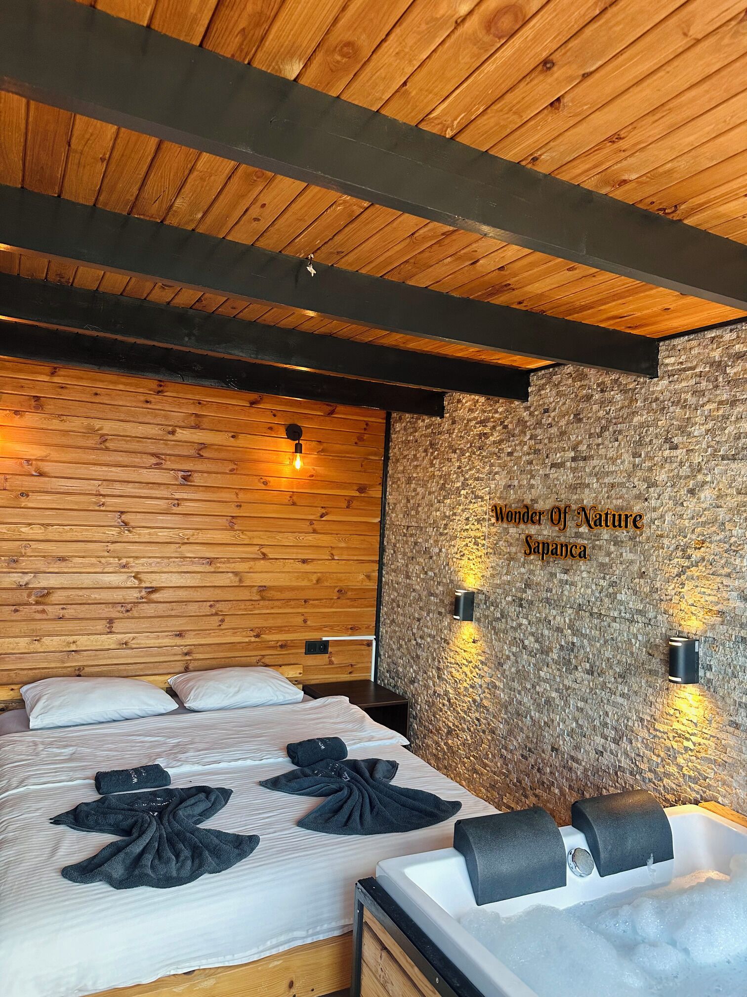A wooden ceiling with exposed beams, a bed with white linens and black towels, a stone wall with a sign, and a bathtub with a black faucet.
