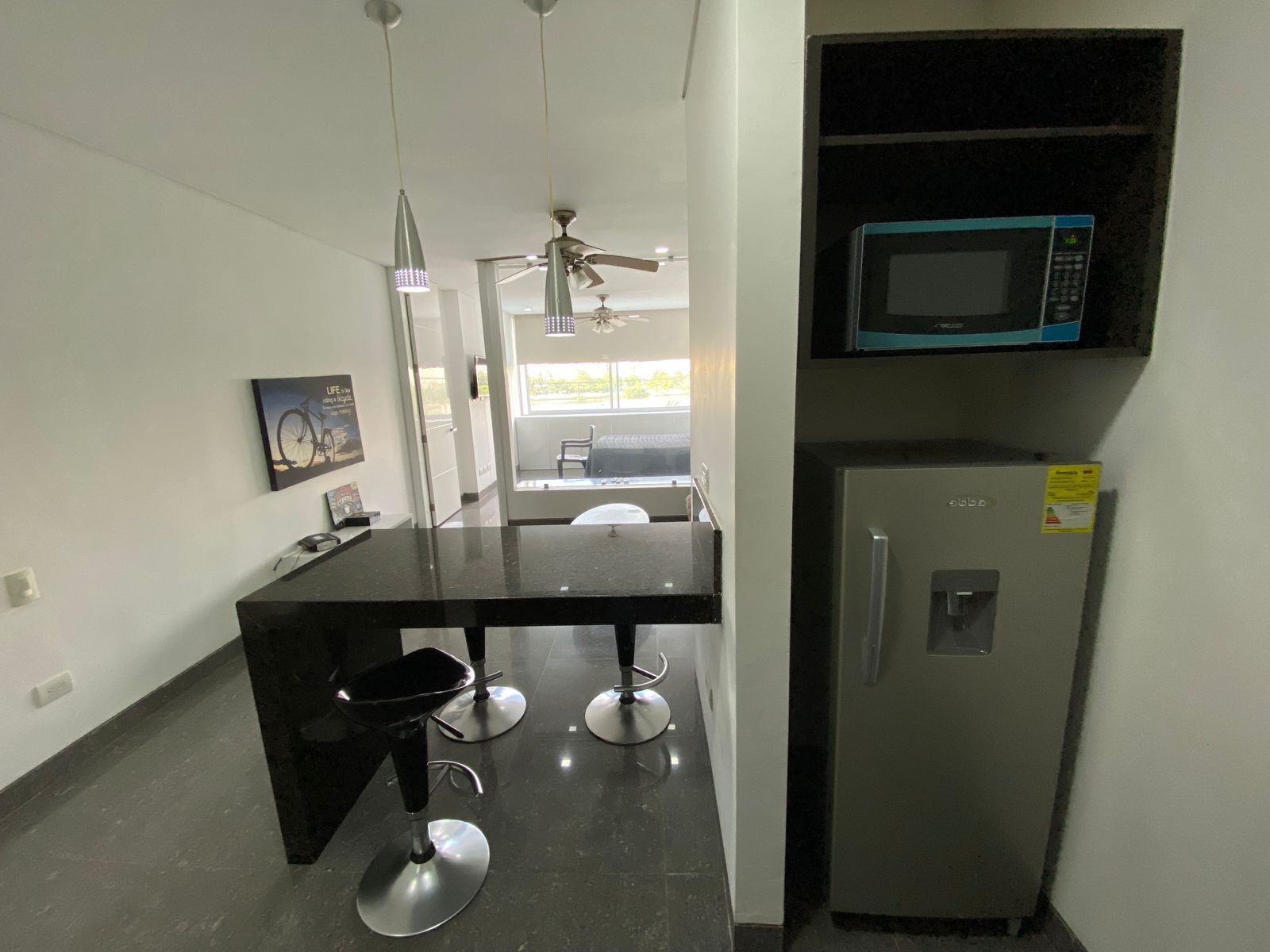 A compact kitchen with a black countertop, a stainless steel refrigerator, a microwave, and a ceiling fan.