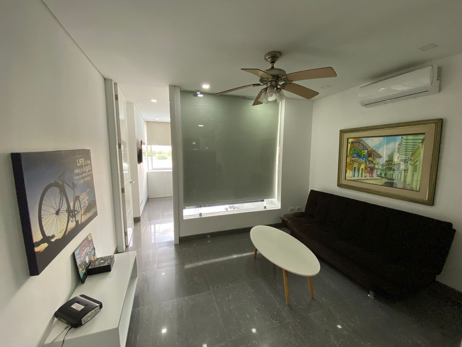 Un salon moderne avec un canapé marron foncé, une table basse blanche et un ventilateur de plafond. On y trouve une grande fenêtre à vitres dépolies, un tableau encadré accroché au mur et une affiche également accrochée au mur.