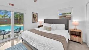 Un dormitorio con cama, ventilador de techo, mesita de noche con lámpara, ventana con vista a una piscina y un cuadro en la pared.