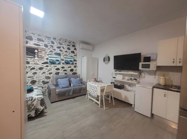 A compact living space with a stone wall, a sofa, a dining table, a TV, and a kitchenette. - Noda House in Tarajalejo (Tarajalejo)