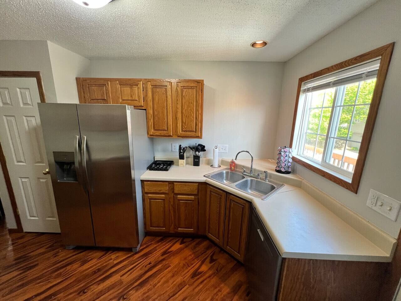 A kitchen with wooden cabinets, a stainless steel refrigerator, a sink, and a window with blinds.
