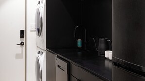 A modern kitchen with dark cabinetry, a built-in washer and dryer, a coffee maker, and a refrigerator. - Coldbrook Bed and Breakfast (Bearsville)