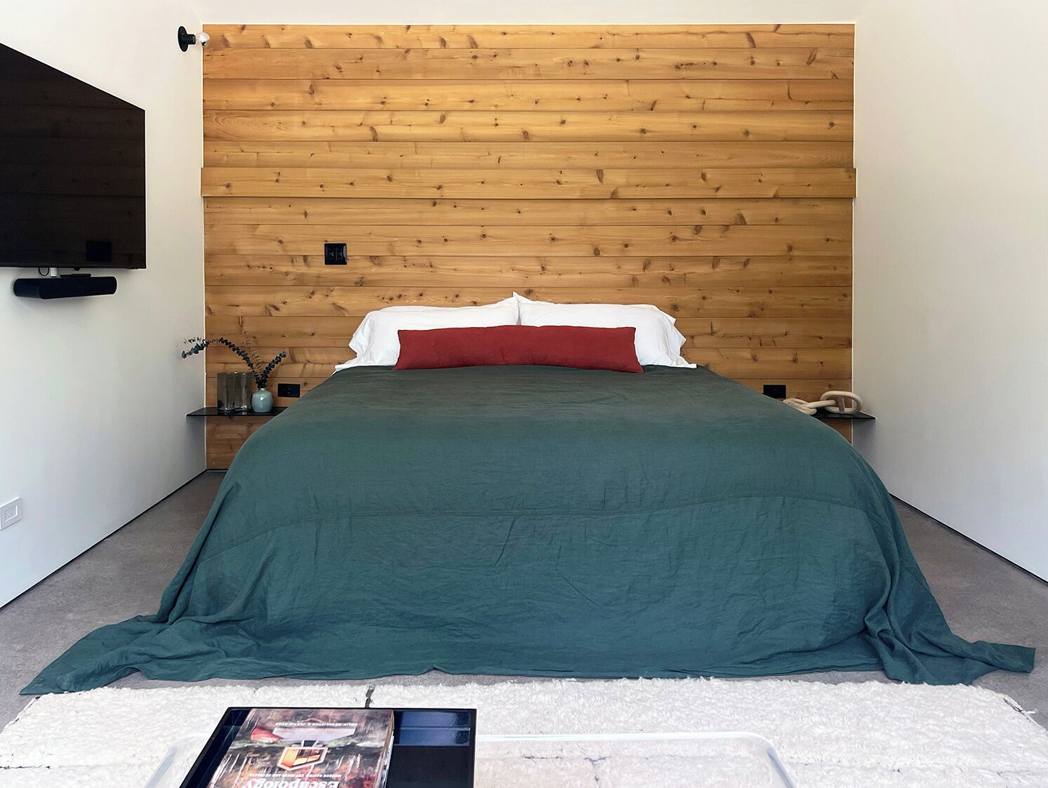 Ein Schlafzimmer mit einem hölzernen Kopfteil, einem Bett mit grün-roter Bettwäsche, einem weißen Teppich und einem kleinen Nachttisch mit einer Lampe.