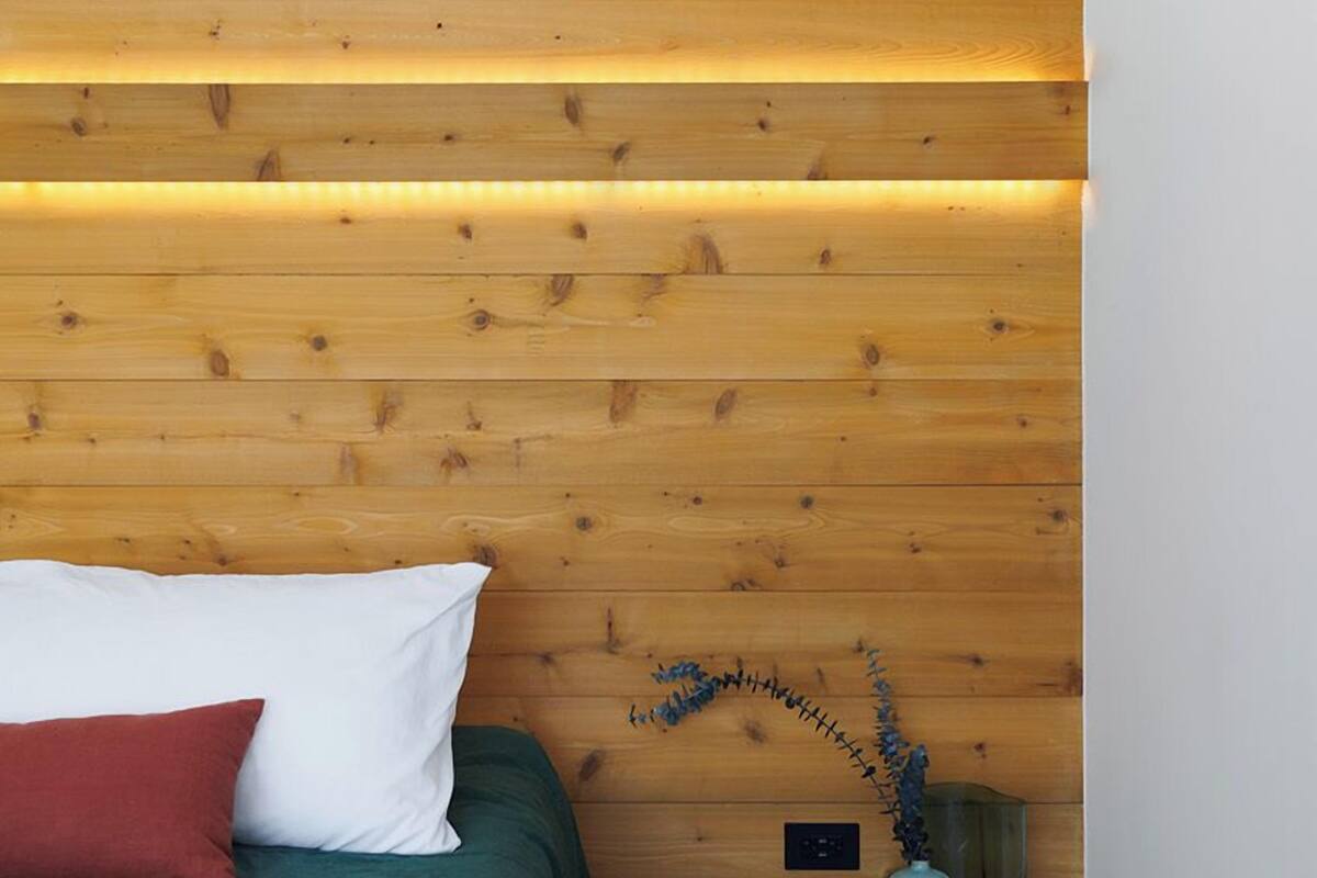 A bedroom with a wooden slat wall, a green bed, a red pillow, and a small nightstand with a vase and a lamp.