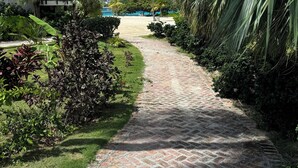 Un camino de ladrillo que lleva a una playa, rodeado de exuberante vegetación y un edificio con balcones.