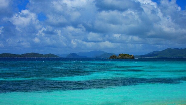 Un paisaje costero con aguas turquesas cristalinas, montañas a lo lejos y una pequeña isla con vegetación.