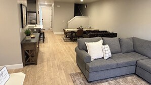 Una sala de estar moderna con un sofá gris, una zona de comedor con mesa y sillas, y una cocina con estufa.