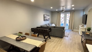 Una sala de estar moderna con sofá, una mesa de comedor con sillas, una mesa de fútbol de mesa y un televisor.