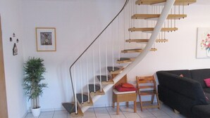 A staircase with wooden steps and white railings, a potted plant, a wicker chair, a black sofa, and a framed picture on the wall. - Haus in Nessmersiel am Wattenmeer (Dornum)