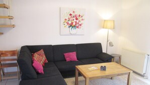 A living room with a dark grey sectional sofa, two pink cushions, a wooden coffee table, a floor lamp, and a painting of flowers on the wall. - Haus in Nessmersiel am Wattenmeer (Dornum)