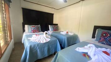 Une chambre d’hôtel avec deux lits, chacun recouvert d’une courtepointe bleue et agrémenté d’une serviette en forme de cygne décoratif. La chambre est équipée d’un ventilateur de plafond et d’une fenêtre avec des rideaux.