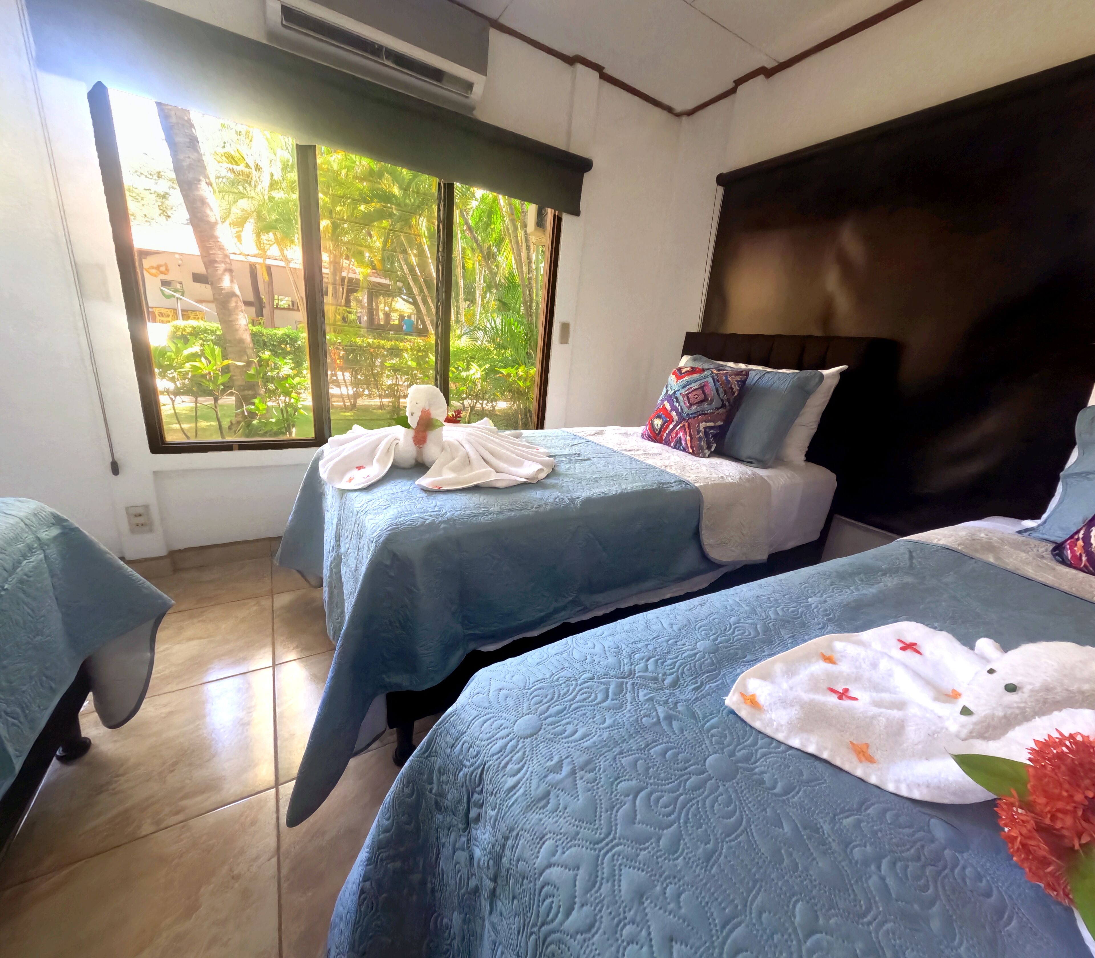 Habitación de hotel con dos camas, cada una con cubrecama azul y toallas blancas. Se ve una ventana con vistas a zonas verdes.