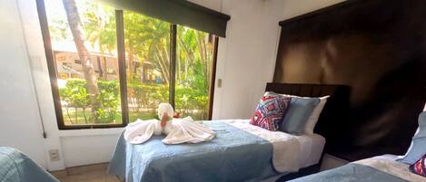 Habitación de hotel con dos camas, cada una con cubrecama azul y toallas blancas. Se ve una ventana con vistas a zonas verdes.