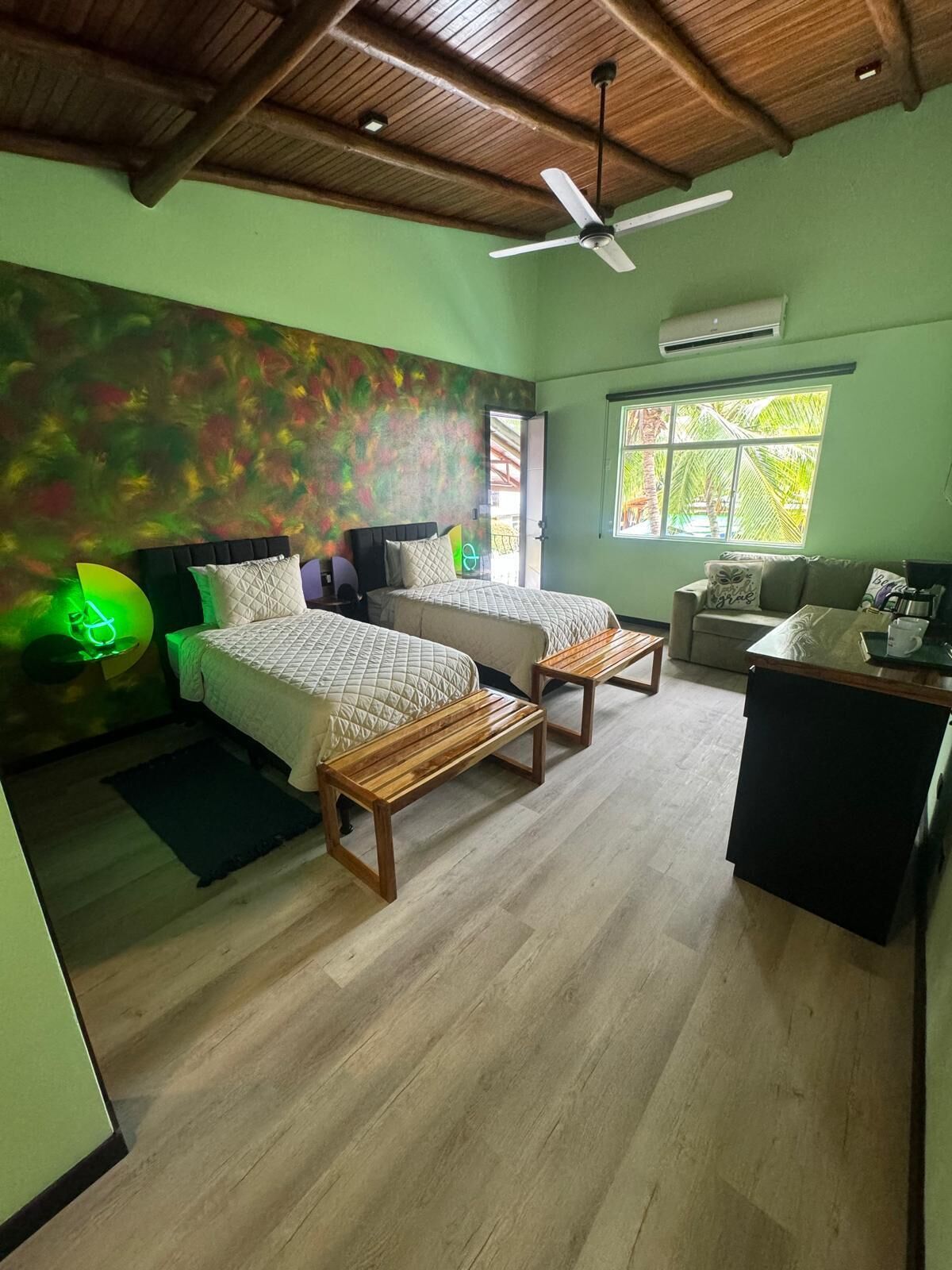A room with two beds, a ceiling fan, a window with a view of palm trees, and a small kitchenette.