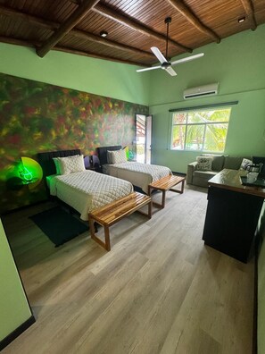 Habitación con dos camas, ventilador de techo, ventana con vista a palmeras y una pequeña kitchenette.