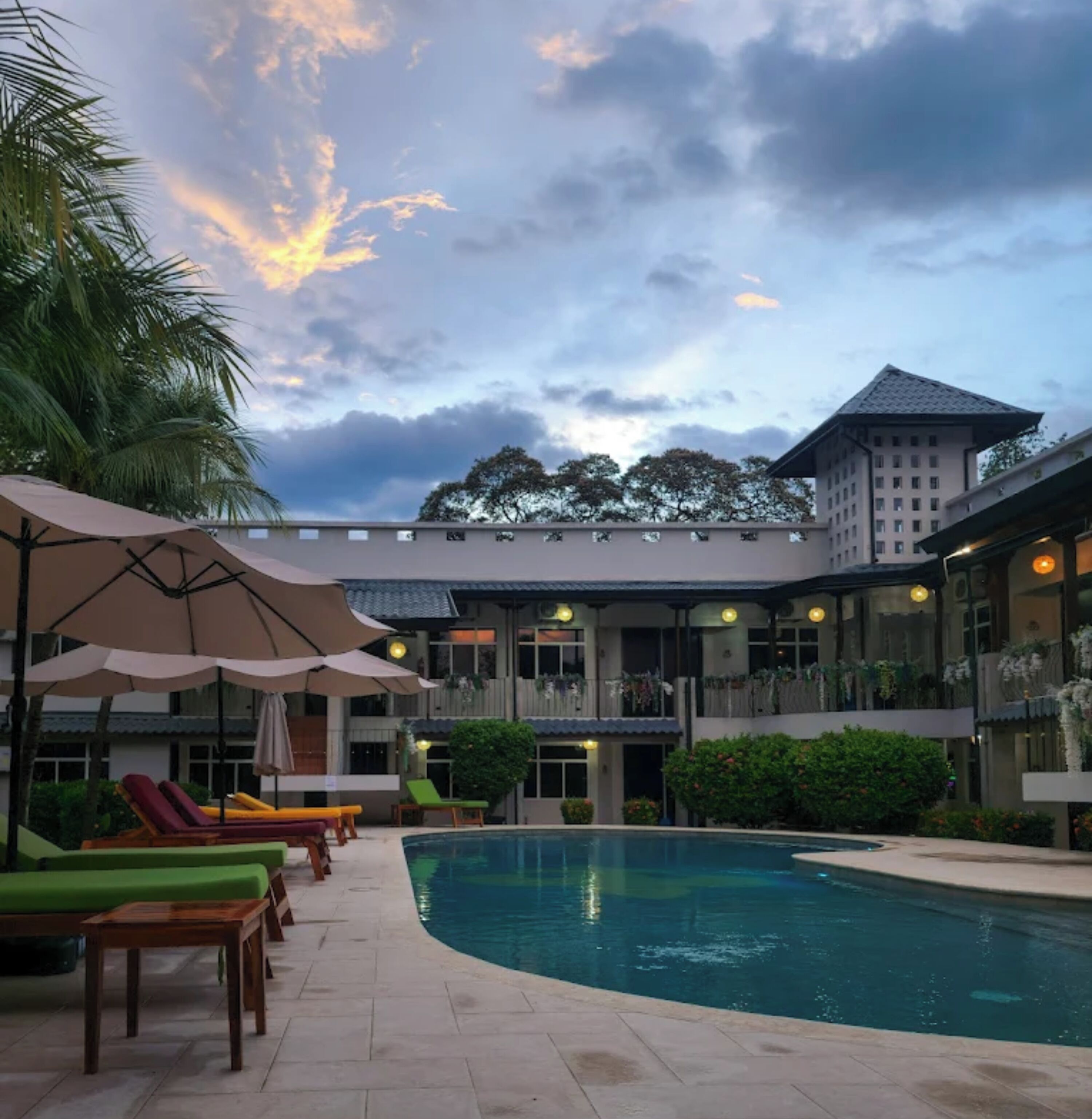 Un espace piscine avec des chaises longues et des parasols, un bâtiment à étages doté de balcons, et un ciel nuageux.
