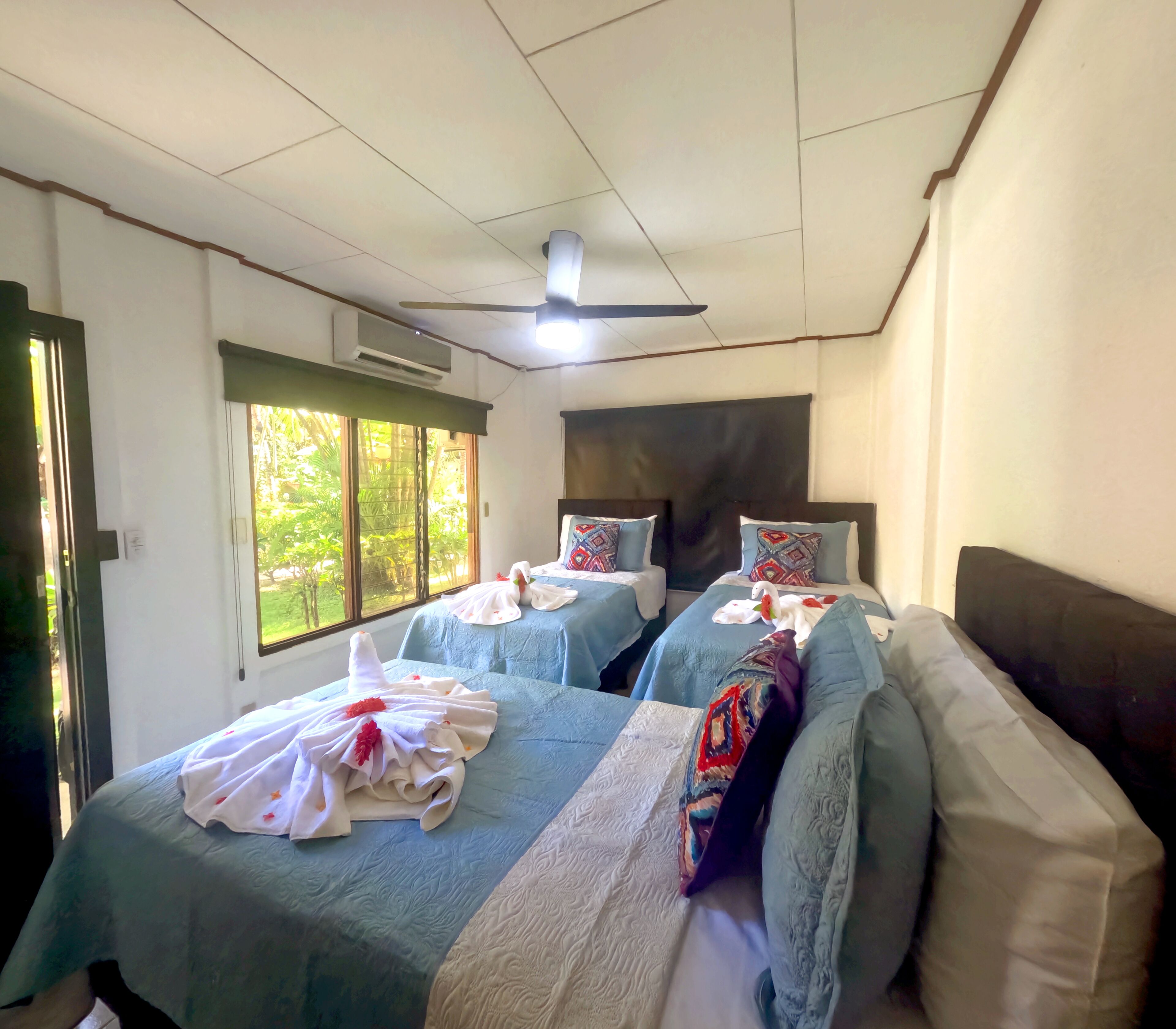 A hotel room with two beds, each with a blue bedspread, white pillows, and a decorative towel. There is a ceiling fan, a window with a view of greenery, and an air conditioning unit on the wall.