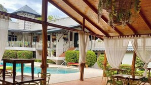 A covered outdoor area with wooden flooring, tables, and chairs, overlooking a swimming pool and a building with a white facade. - Parti Gras Hotel & Villas en Potrero (Tempate)