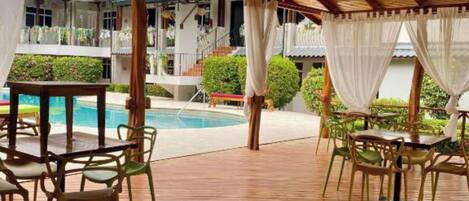A covered outdoor area with wooden flooring, tables, and chairs, overlooking a swimming pool and a building with a white facade.