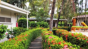 A pathway flanked by neatly trimmed hedges and vibrant flowers, leading to a building with a white exterior and a covered outdoor seating area. - Parti Gras Hotel & Villas en Potrero (Tempate)