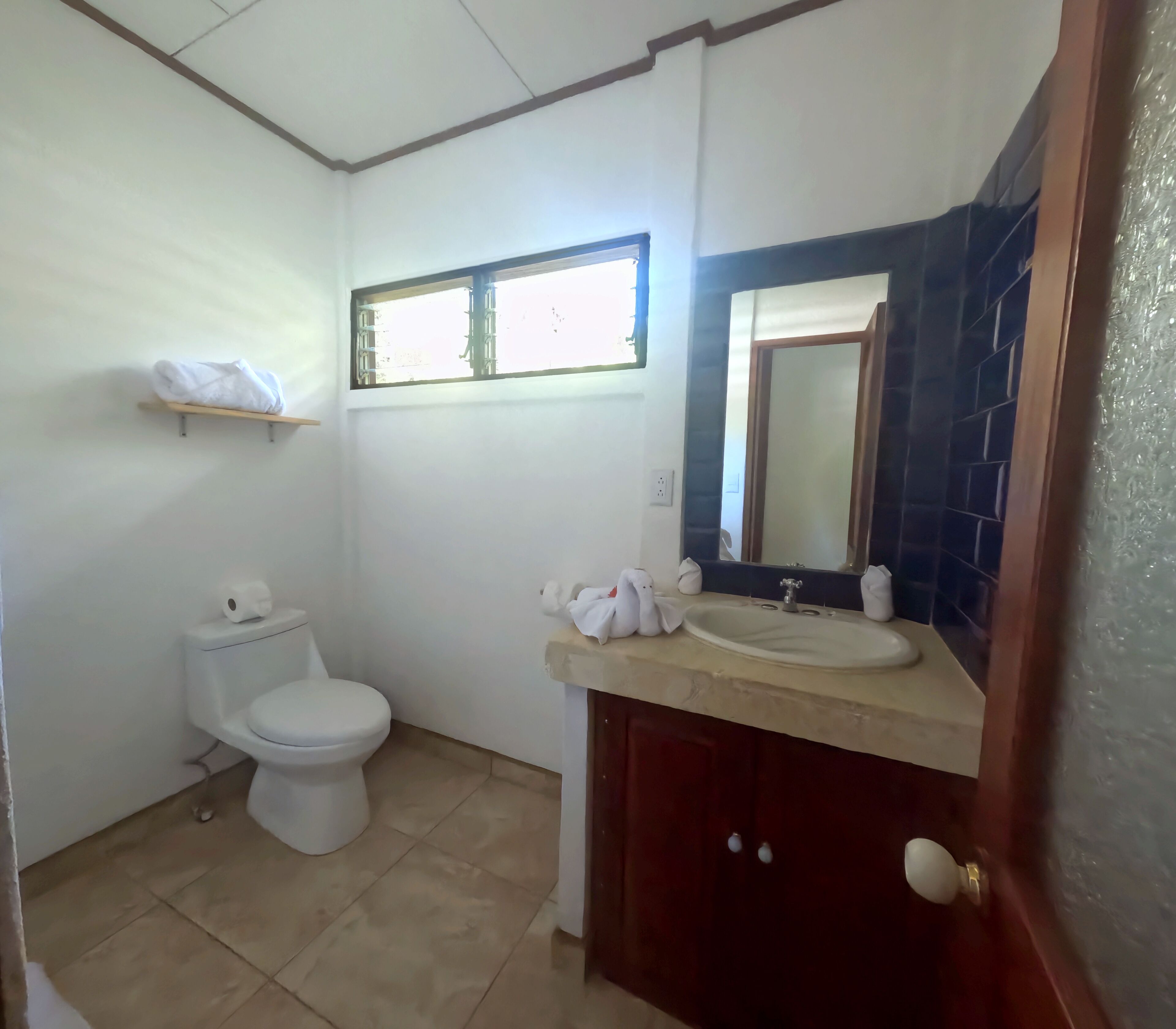 Un baño pequeño con inodoro, lavamanos y espejo. Hay una ventana sobre el inodoro y un estante con toallas.