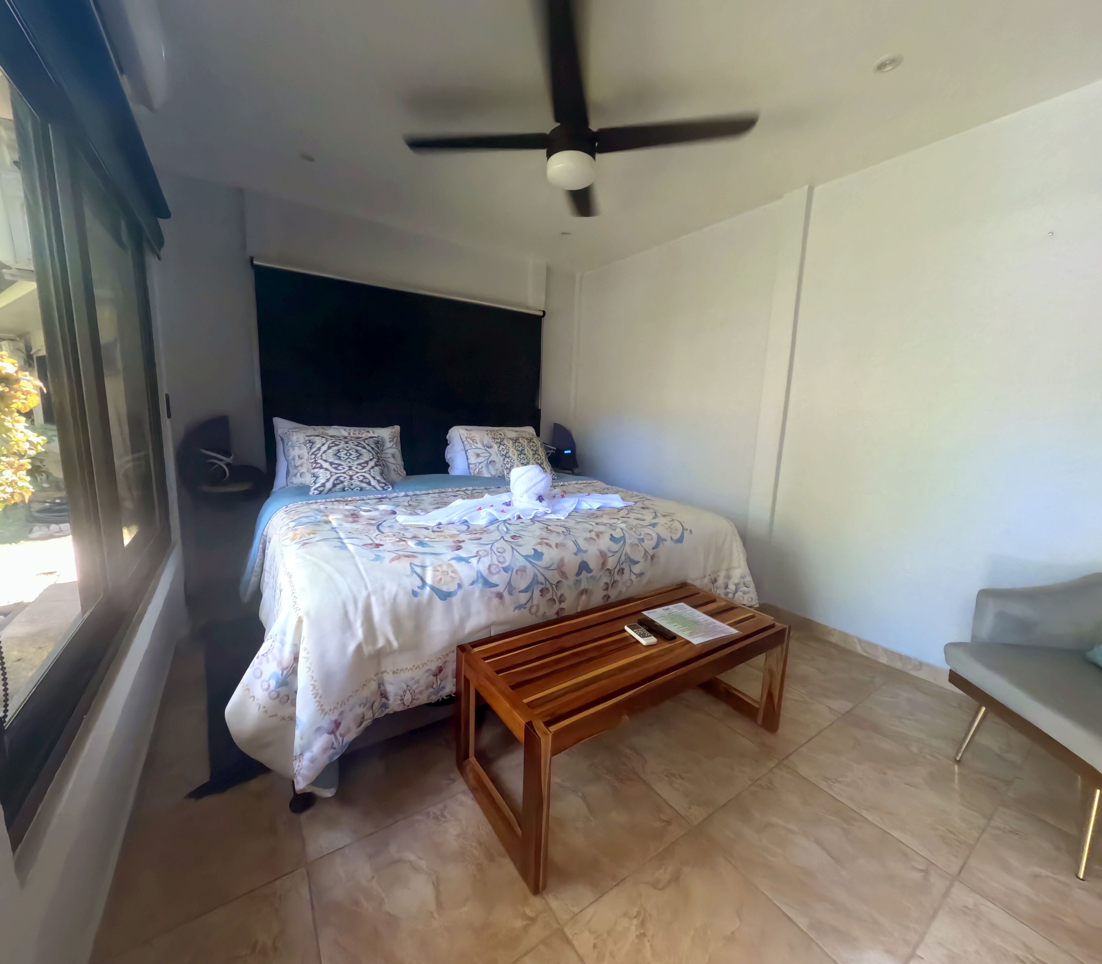 A bedroom with a bed, a wooden bedside table, a ceiling fan, and a window.