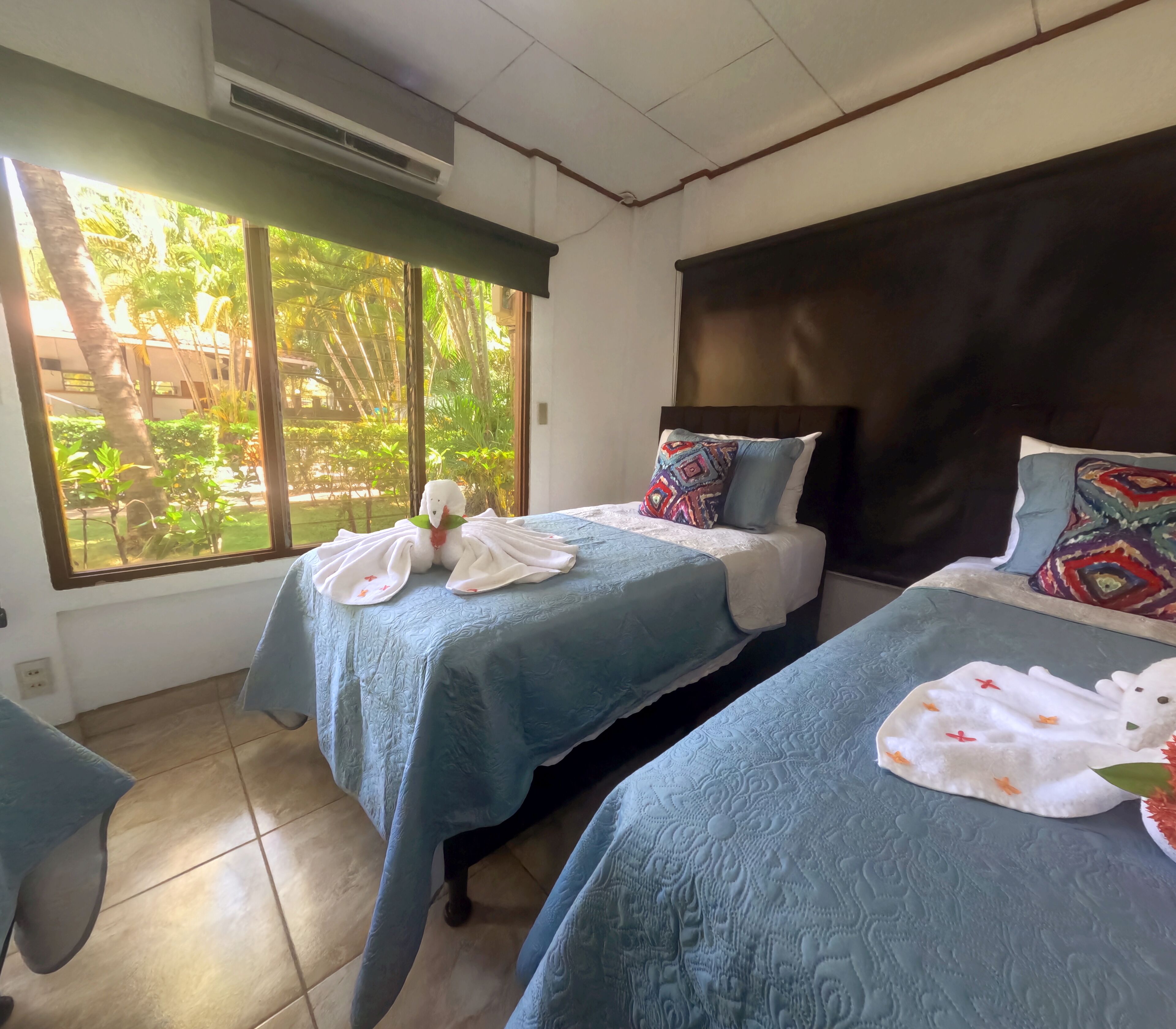 Une chambre d’hôtel avec deux lits, chacun recouvert d’une couverture bleue et agrémenté d’oreillers. Une fenêtre offre une vue sur la verdure environnante.