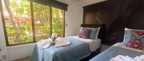 Une chambre d’hôtel avec deux lits, chacun recouvert d’une couverture bleue et agrémenté d’oreillers. Une fenêtre offre une vue sur la verdure environnante.