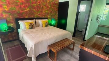 Un dormitorio con cama, mesitas de noche, un banco, techo de madera y un baño visible a través de una puerta abierta.
