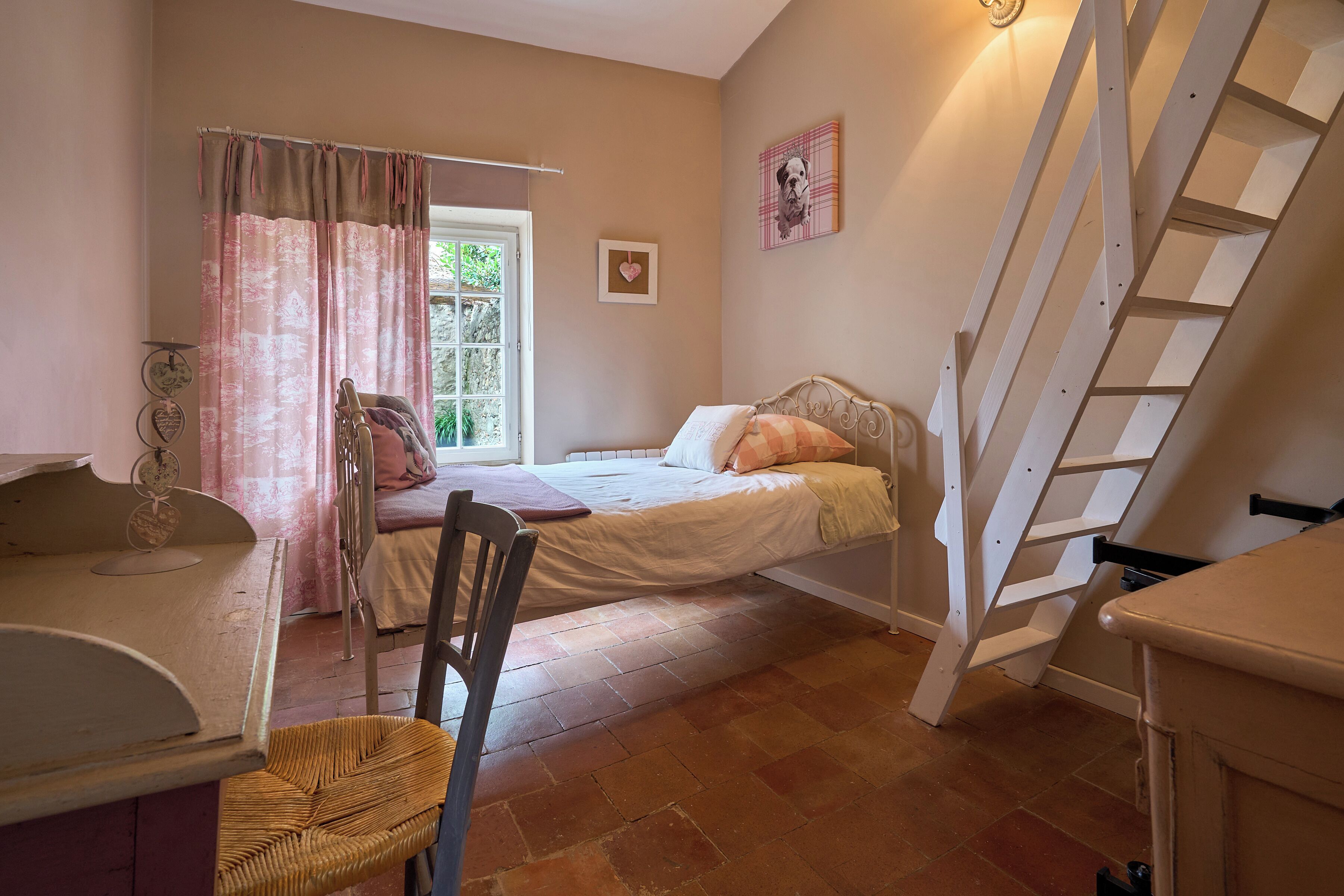 A bedroom with a bed, a chair, a desk, a ladder, and a window with curtains.