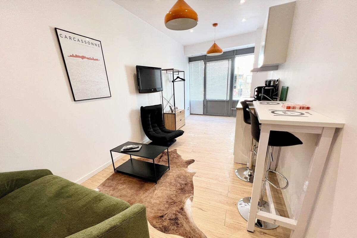 Una sala moderna con un sofá verde, un sillón negro, una mesita negra y un televisor montado. Se ve un área de cocina con encimera blanca y taburetes de bar al fondo.