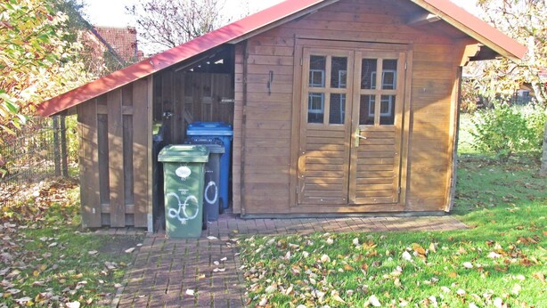 A wooden shed with a red roof, a door, and two windows. There are two green trash bins in front of the shed. - Allergiker Freundliches Haus in Nessmersiel (Dornum)
