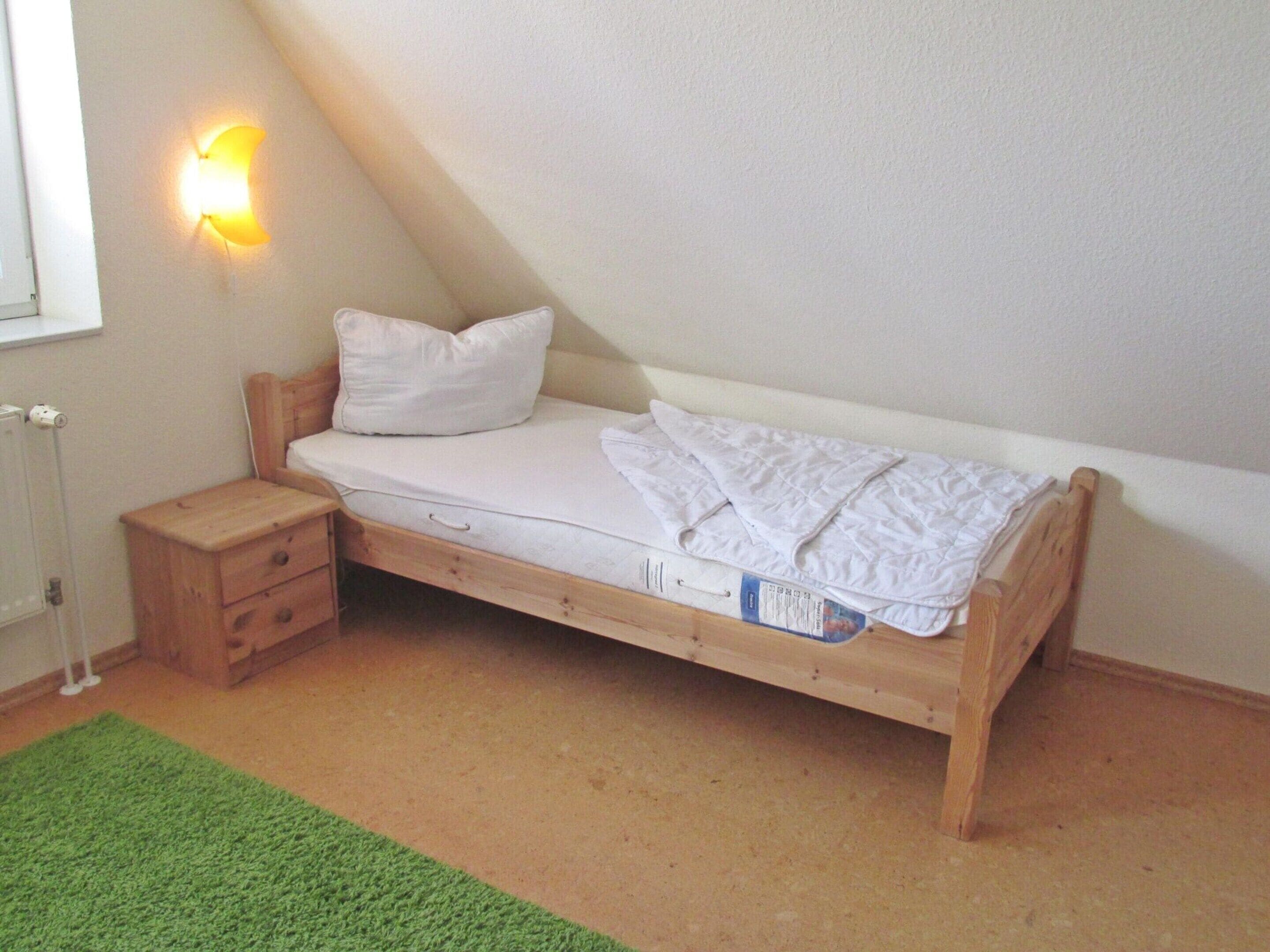 A single bed with a wooden frame, a small bedside table, and a green rug.