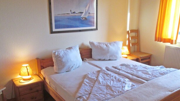A neatly made bed with white linens, two bedside tables with lamps, and a painting of a sailboat on the wall. - Allergiker Freundliches Haus in Nessmersiel (Dornum)