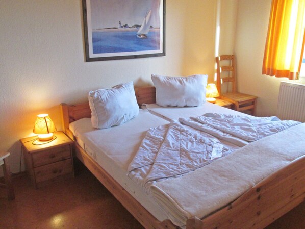 A neatly made bed with white linens, two bedside tables with lamps, and a painting of a sailboat on the wall. - Allergiker Freundliches Haus in Nessmersiel (Dornum)