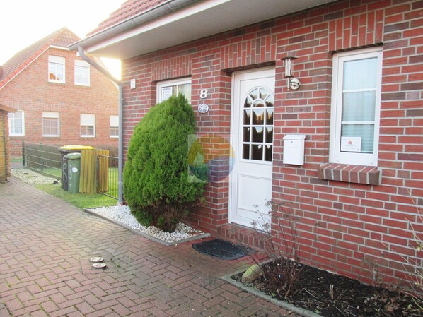 A red brick building with a white door and windows, a small garden area with a hedge, and a metal fence in the background.