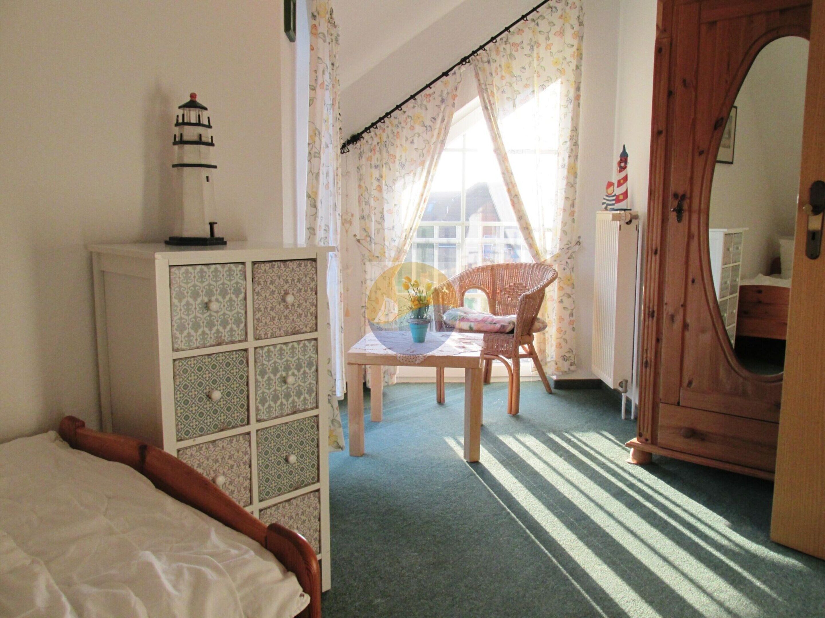 Een kamer met een bed, een klein kastje, een tafel met een vaas, een rieten stoel en een houten garderobe.