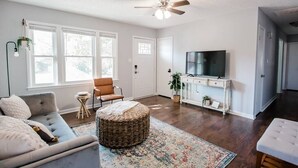 A living room with a sofa, ottoman, and a television on a white cabinet. - HOT TUB, Fenced in Yard, Pet Friendly, Game room (Fayetteville)