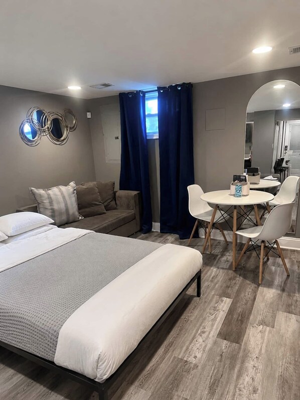 A hotel room with a large bed, a sofa, a dining table with chairs, a mirror, and a window with curtains.