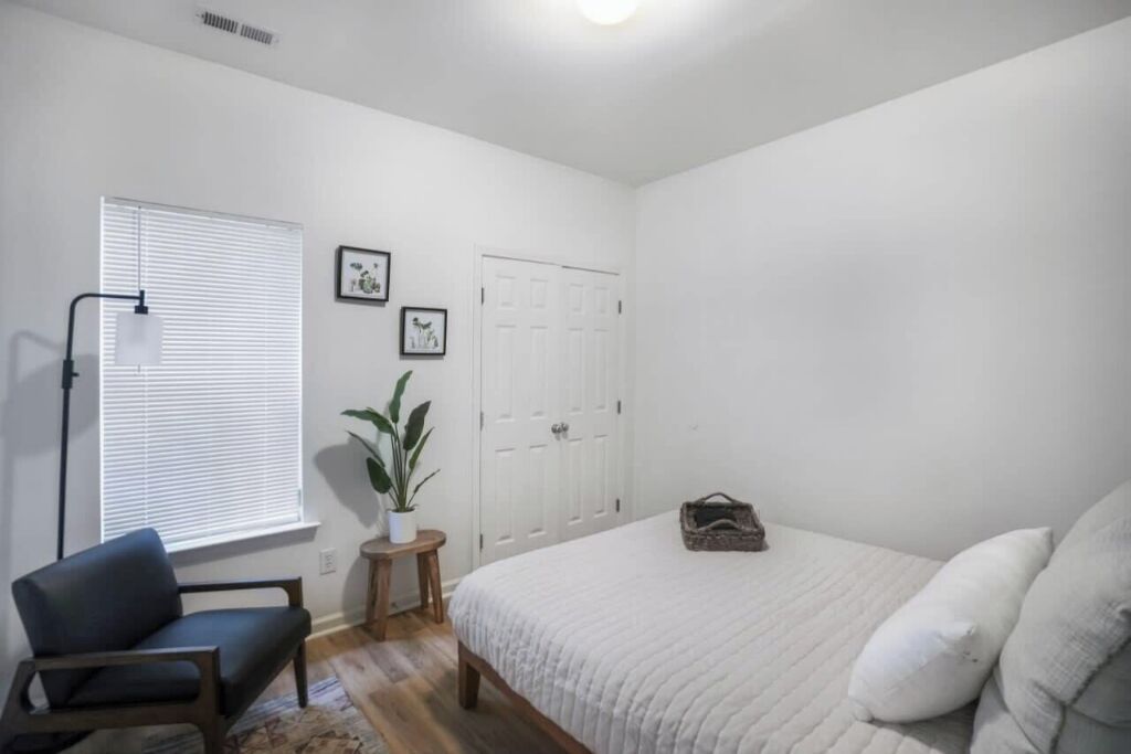 Een slaapkamer met een bed, een stoel, een klein tafeltje met een plant en een deur.