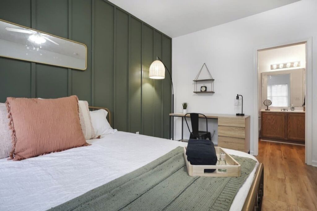 A bedroom with a bed, bedside table, dresser, and a small desk with a chair. There is a mirror above the dresser and a shelf with a clock.