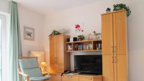 A living room with a sofa, a TV, wooden cabinets, and a bookshelf. - Haus in Ostfriesland mit Garten (Dornum)