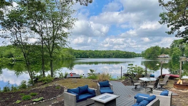 A deck with wicker furniture and a table, overlooking a lake with a house in the background.